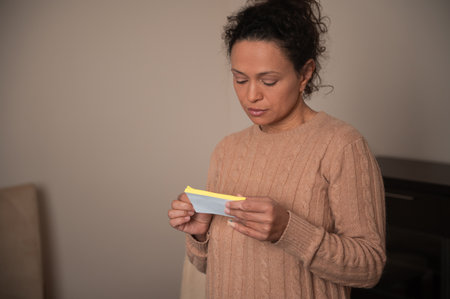 A woman stands indoors in a cozy sweater, attentively reading a note. The setting is warm and inviting, conveying a sense of quiet contemplation.の写真素材