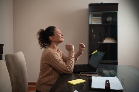 A woman sits at a table, cheering happily while looking at her laptop, symbolizing accomplishment.