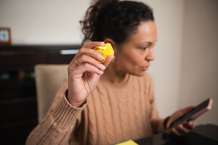 A woman in a brown sweater holds a crumpled yellow paper, focusing intently on her smartphone at a...