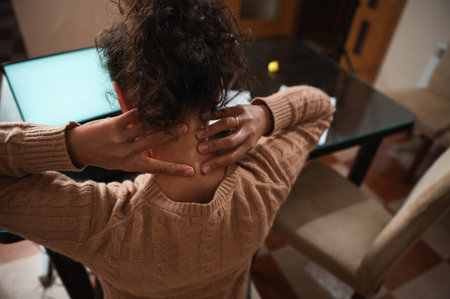 A woman sitting at a glass table massages her neck with both hands, seeking relief from tension or stiffness. Her cozy sweater suggests a relaxed environment, possibly a home or office setting.の写真素材