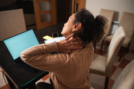 A woman stretches her neck while sitting at a desk, working from home. The scene captures the need for a break during long hours of computer use.の写真素材