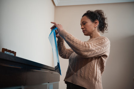 A woman stands in a cozy room, examining a document, focused and thoughtful. Her casual attire and serene environment suggest she is in a comfortable, private setting.の写真素材