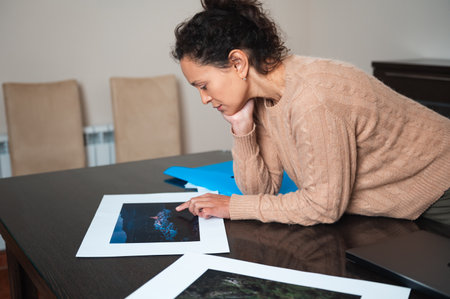A woman carefully studies photographs laid out on a table, showcasing attention to detail and focused engagement in a calm, indoor environment.の写真素材