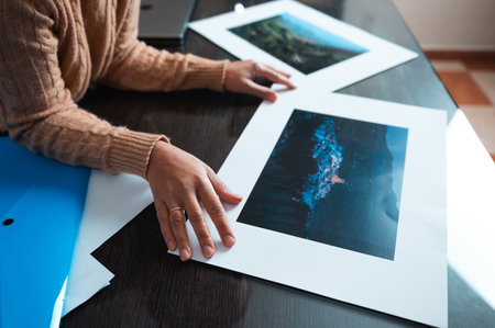 Hands of a person analyzing photographic prints on a wooden table, reflecting careful consideration of the images displayed.の写真素材