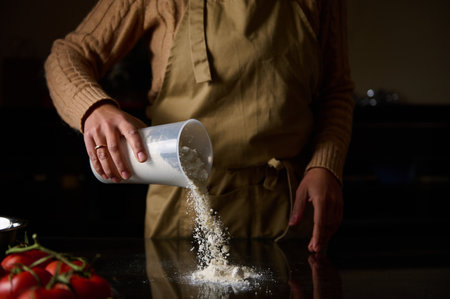A person in an apron sifts flour onto a kitchen counter, ready for baking. Fresh tomatoes sit...