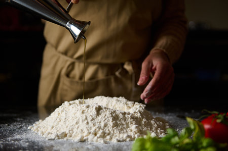 Close-up of a chef in an apron pouring oil onto a mound of flour, beginning the process of making fresh dough. Fresh ingredients are arranged in the foreground, indicating a vibrant and natural culinary experience.の写真素材