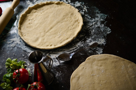 Top view of homemade pizza dough preparation with fresh basil, tomatoes, and kitchen utensils on a floured surface, highlighting the rustic cooking ambiance.の写真素材