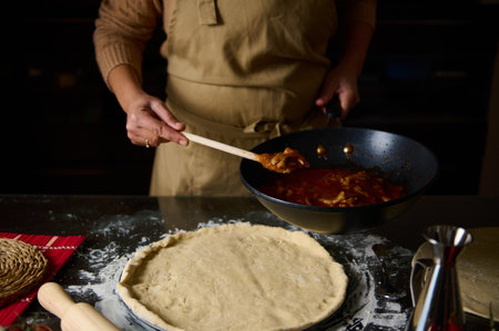 A woman in an apron prepares homemade pizza, spreading sauce over the dough on a floured surface.