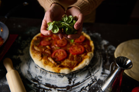 A close-up of hands placing fresh basil on a homemade pizza topped with tomatoes, showcasing cooking preparation.の写真素材