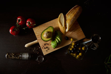 Top view of fresh ingredients including avocado, tomatoes, olives, bread, and spices on a wooden board.の写真素材