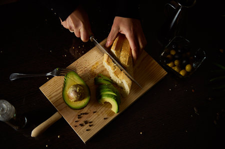 A person slices bread and avocado on a wooden board in a dimly lit setting, creating a rustic and minimalistic atmosphere.の写真素材