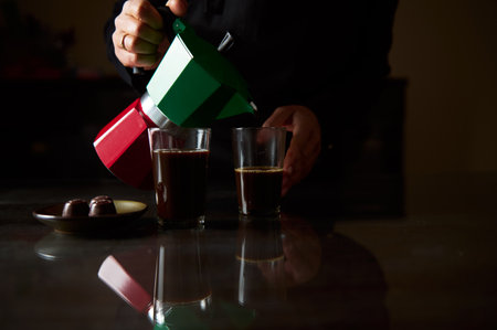 A person is pouring freshly brewed coffee from a moka pot into glass cups. Dark tones create a cozy...