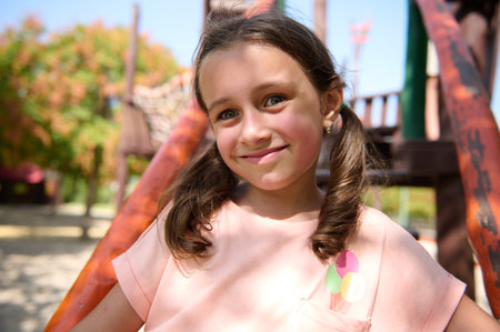 A cheerful young girl with pigtails smiles brightly while playing at a sunny playground.