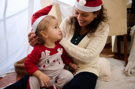 A joyful mother and child share a festive moment indoors, both wearing Santa hats, creating warm...