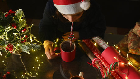 A child in a Santa hat drinks from a mug amidst Christmas lights, greenery, and wrapped presents,...
