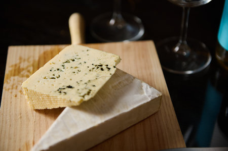A display of two types of cheese resting on a wooden board, accompanied by wine glasses. The image evokes a rustic atmosphere, perfect for gourmet and culinary themes.の写真素材