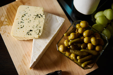 A selection of cheese, pickles, and grapes placed elegantly on a wooden board. Perfect for dining, snacks, and gourmet presentations.の写真素材
