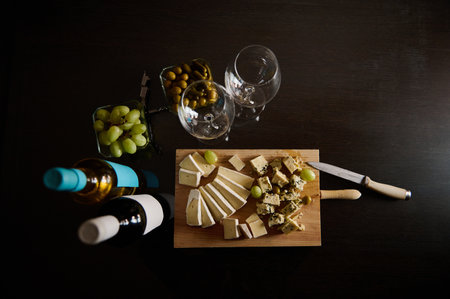 A sophisticated table arrangement featuring assorted cheese, wine bottles, glassware, and garnishes on dark wood.の写真素材