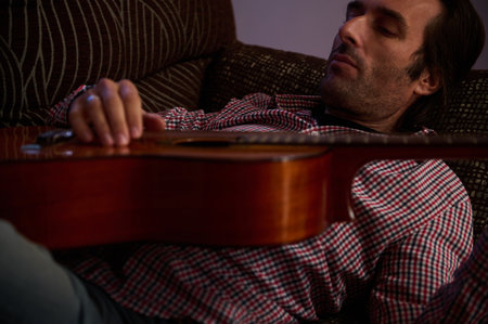 A man dressed casually is lying on a couch with a guitar, capturing relaxation and a love for music...