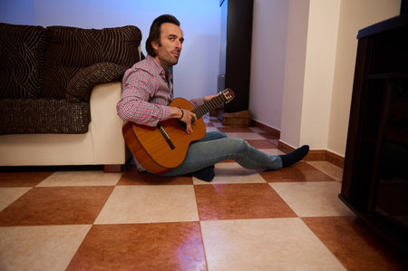 A man in casual attire playing an acoustic guitar, sitting on the floor in a cozy living room, embracing creativity and relaxation. The tiled flooring and furniture create a homey atmosphere.の写真素材