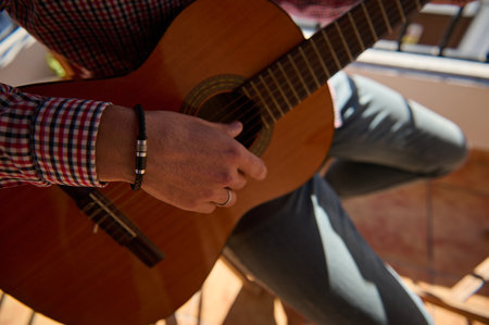 A close-up view of a person in casual clothing playing a wooden acoustic guitar during daytime,...