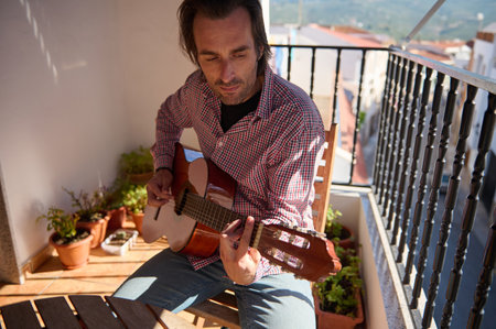 A person enjoys playing an acoustic guitar outside on a sunny terrace surrounded by potted plants, creating a relaxed and musical ambiance.の写真素材