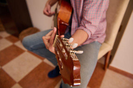 A musician adjusts the tuning pegs of an acoustic guitar while seated at home. Captured in a cozy indoor setting with details enhancing the activity of fine-tuning his instrument.の写真素材