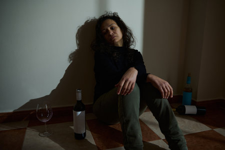 A woman is sitting on the floor in a dimly lit room, surrounded by wine bottles, reflecting distress and struggles with emotions.の写真素材