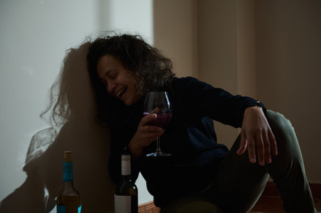 A woman seated on the floor holding a glass of wine, expressing relaxation and contentment, illuminated by soft light casting shadows on the wall.の写真素材