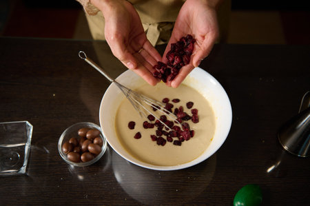A person in a kitchen pours dried fruit into a bowl of batter while preparing a dessert.