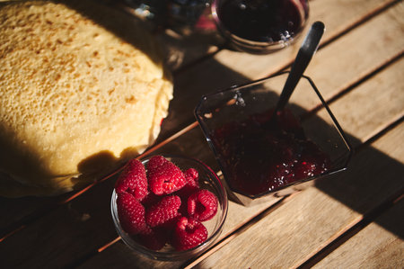 Pancakes and fresh raspberries with jam in bowls on a wooden table in sunlight, perfect for breakfast or dessert ideas.の写真素材