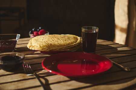 A stack of golden crepes sits on a rustic wooden table accompanied by drinks, jam, and fresh berries. A casual and inviting meal setup reflecting a warm homely atmosphere.の写真素材