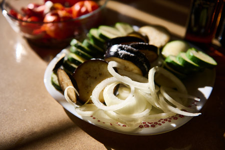 A plate of diverse freshly cut vegetables including cucumber, onion, and eggplant displayed in soft sunlight.の写真素材