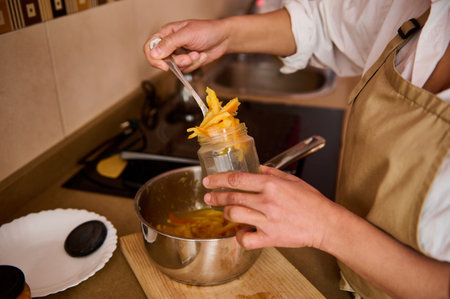 A person wearing an apron fills a glass jar with preserved fruit in a kitchen. A saucepan and a...