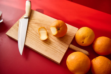 Fresh whole and sliced oranges displayed on a wooden cutting board with a kitchen knife, placed atop a red surface, showcasing vibrant colors and a kitchen preparation scene.の写真素材