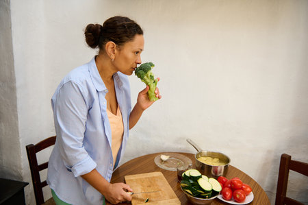 A woman carefully preparing fresh vegetables, including broccoli and zucchini, in a cozy kitchen area. The setting emphasizes home cooking with a focus on healthy eating and natural ingredients.の写真素材