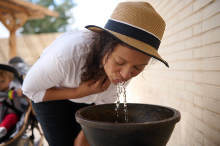 A person wearing a hat drinks water from a fountain in a sunny outdoor setting, looking refreshed and content.の写真素材