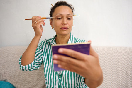 A woman sits on a couch, using a brush and mirror to apply makeup. The relaxed atmosphere emphasizes personal care and routine. She is wearing a striped green shirt, looking focused on her task.の写真素材