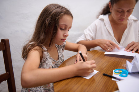 A young girl focuses on her craft project alongside her mother at a table.の写真素材