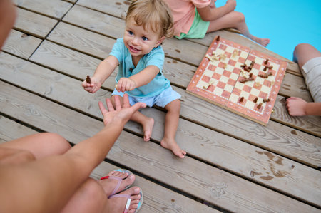 A young child hands a chess piece to an adult, sitting on a wooden deck near a pool, alongside a chessboard and family activities.の写真素材