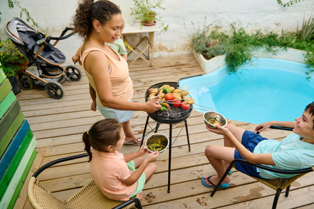 A family gathers around a backyard barbecue by the pool, sharing food and happiness. The scene radiates togetherness, relaxation, and summer vibes on a beautiful sunny day.の写真素材