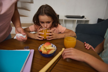 A little girl enjoys a glass of orange juice sitting at a table with family members, surrounded by healthy snacks and colorful details, embodying family life, balanced eating, and home comfort.の写真素材