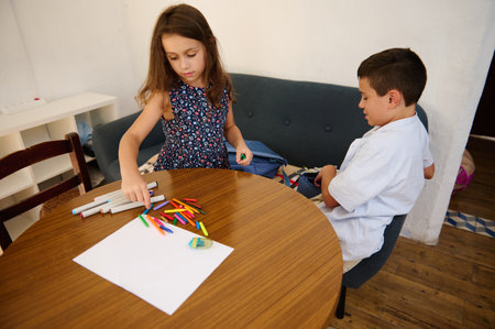 Two children sit at a wooden table working on creative crafts with markers, pencils, and paper. The...