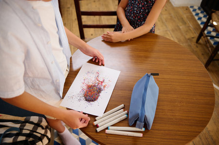 Children seated at a wooden table sharing and discussing a creative artwork, with colorful markers...