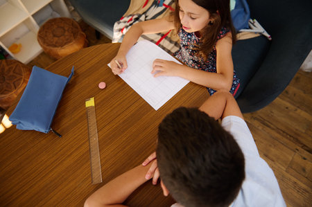 A young girl and boy working on homework together at a wooden table in a colorful classroom setting.