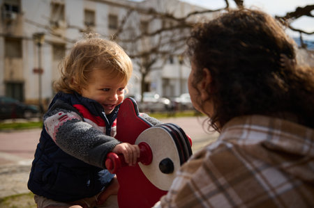 A young child rides a red playground toy, smiling while a mother watches nearby, enjoying outdoor bonding time in a warm and sunny urban park setting.の写真素材