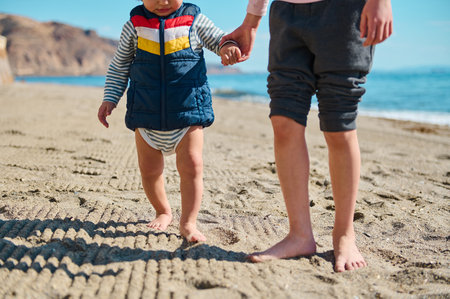 A child holding hands is walking barefoot on a sunlit beach during summer, capturing a warm and...