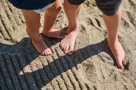 Barefoot feet of a child and an adult standing on sandy ground, featuring shadows and textured...