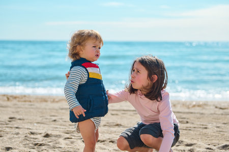 A toddler and older child enjoy their time on a peaceful sandy beach, engaging in curiosity,...