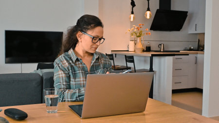 A woman is focused on remote work at her laptop in a stylish and modern home, encompassing...